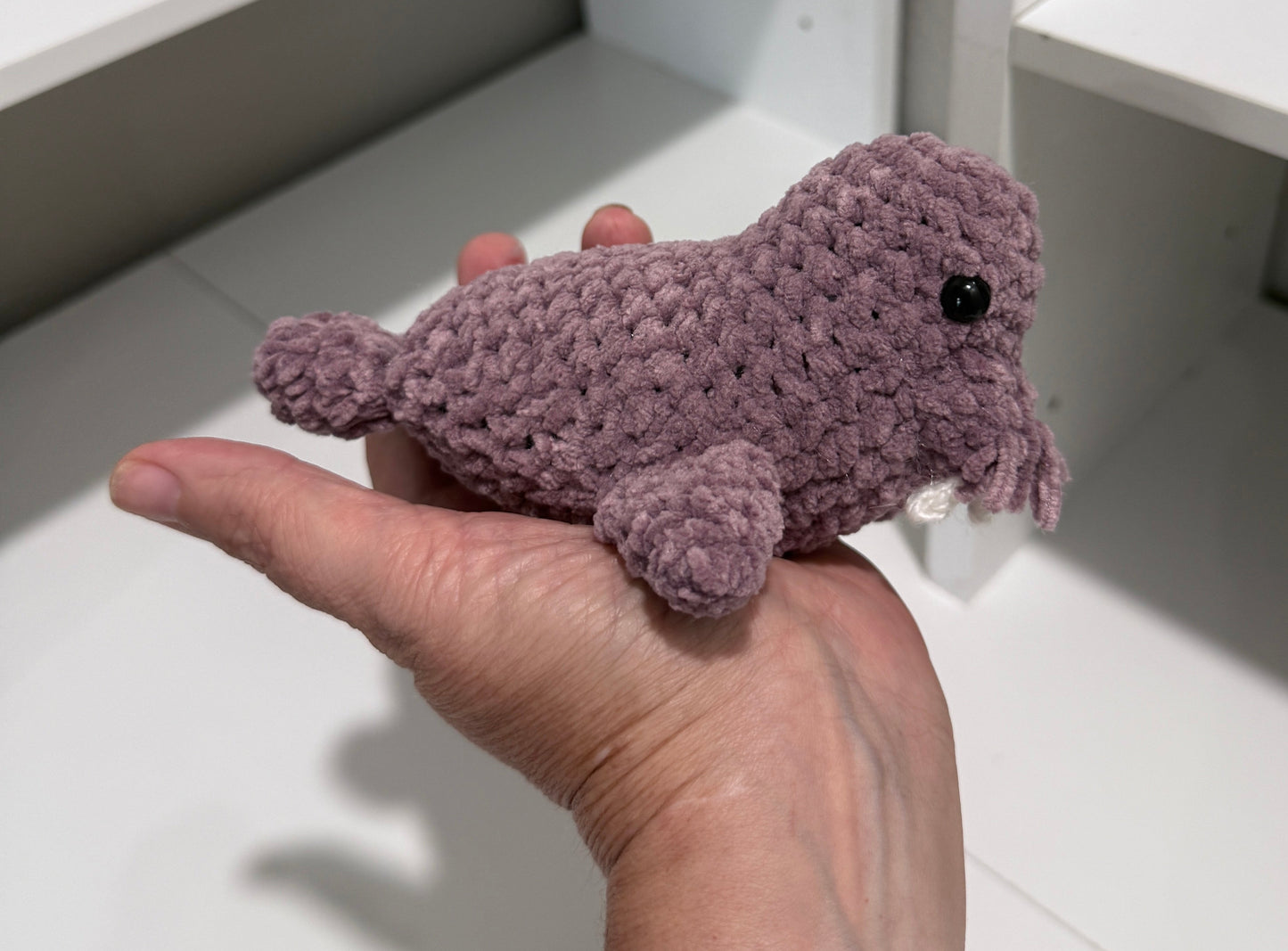 Hand holding a palm size dusty lavender crocheted walrus on a white surface