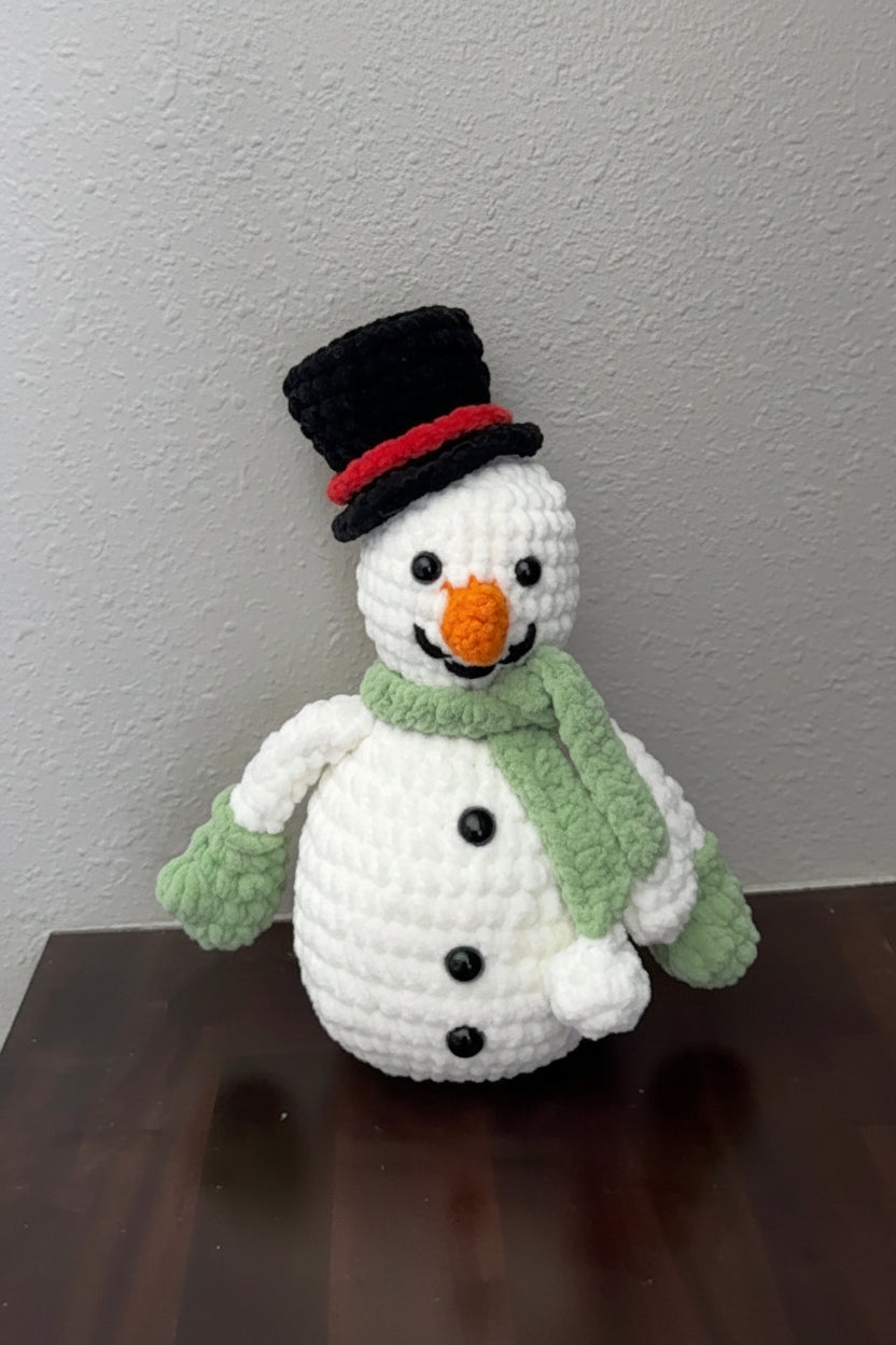 Crocheted snowman with a black top hat, orange carrot nose, and green scarf on a wooden surface.