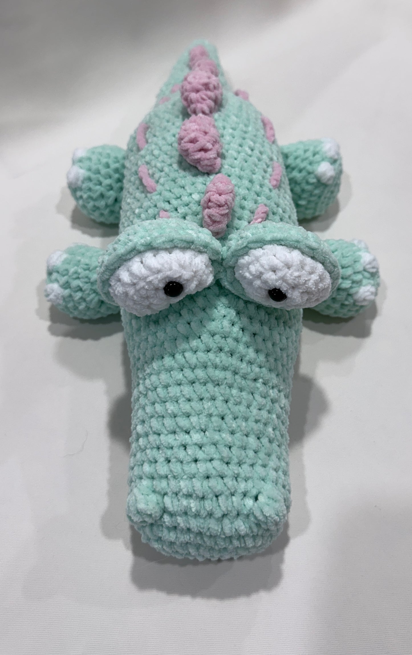 Crocheted crocodile with a mint green body, white eyes, and pink accents on a white background