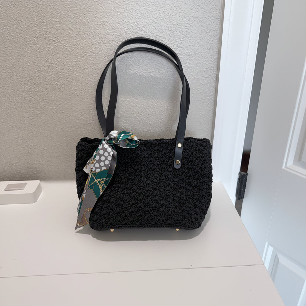 Black crochet handbag with a decorative ribbon held against a white wall.