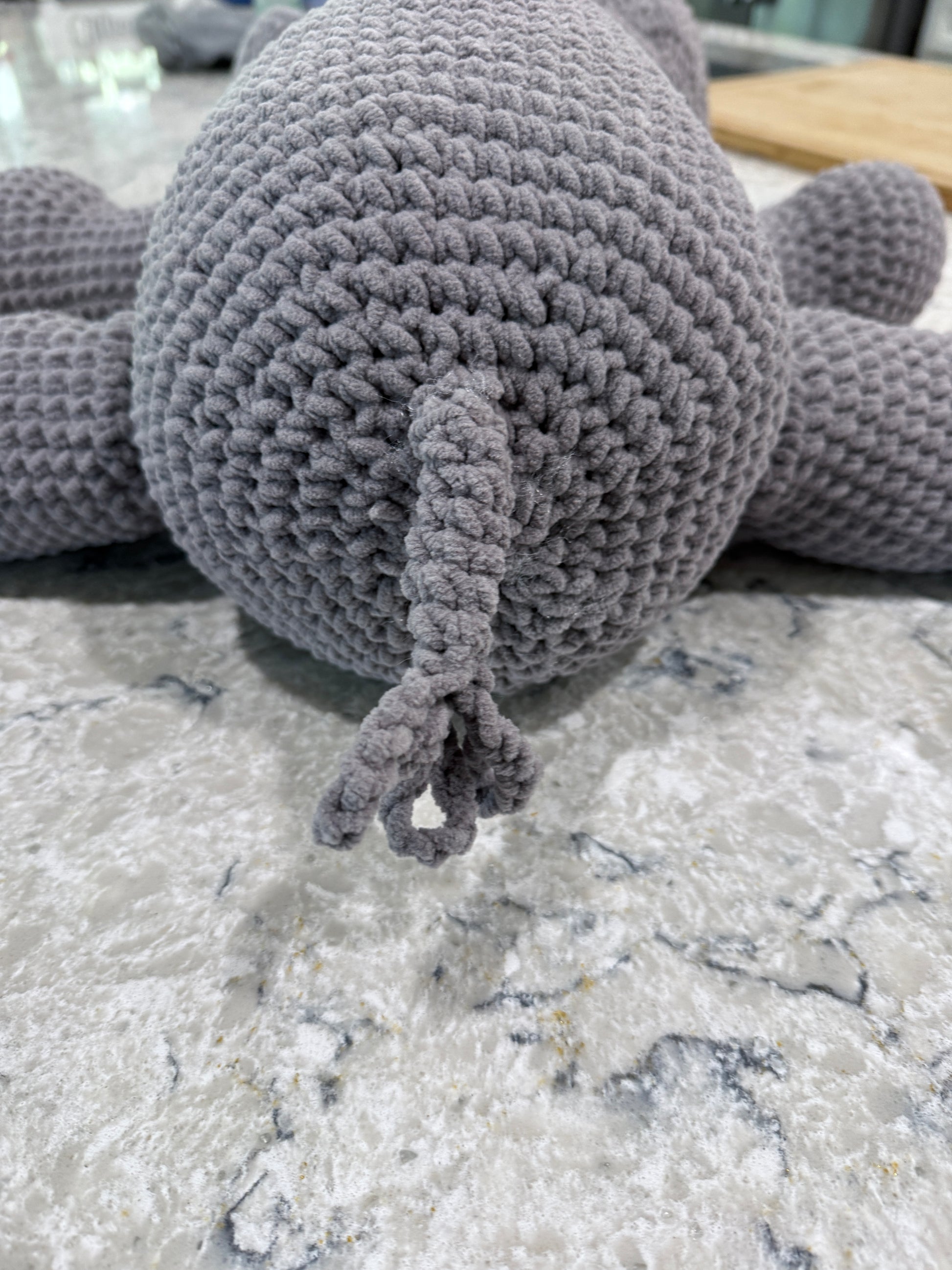A close-up of a gray crocheted plush toy, showing the back and tail, resting on a gray and white marble surface.