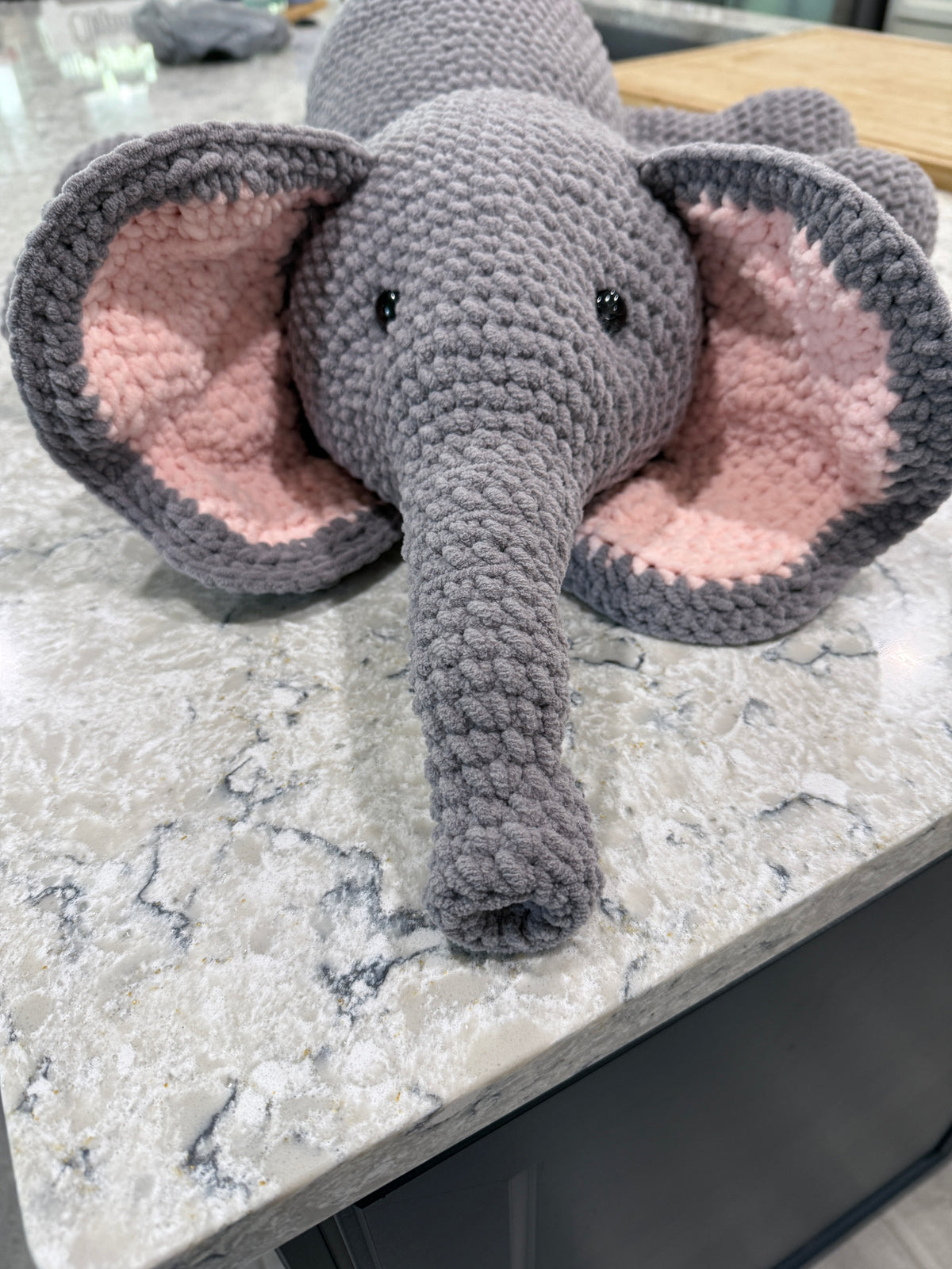A crocheted plush elephant toy with a gray body, long trunk, and large pink inner ears.