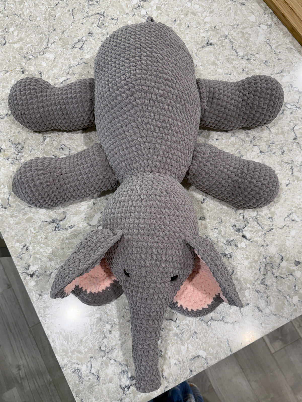 A crocheted elephant plush toy in medium gray with pink inner ears, lying on a light gray marble-patterned countertop.