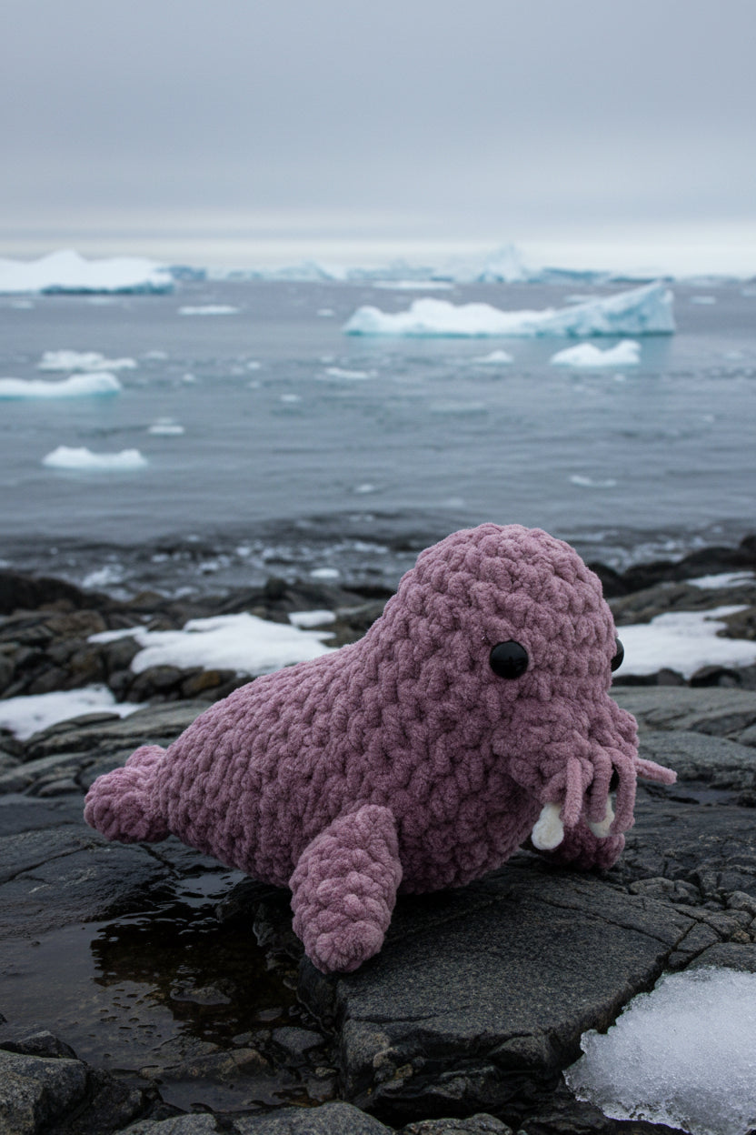 Crocheted dusty lavender walrus on a rocky shore with icebergs in the background