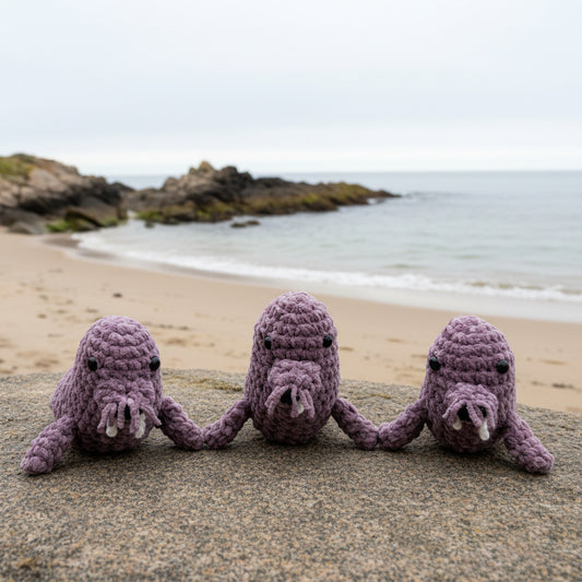 Three dusty lavender crochet walrus sitting on a white surface with a neutral background.