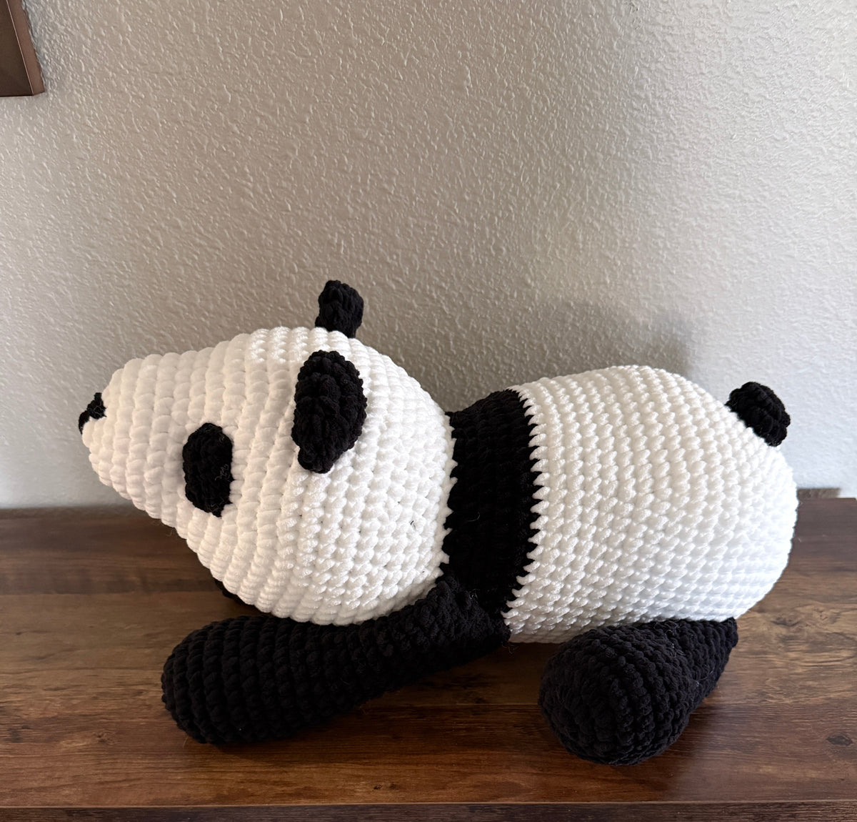 Side view of a crocheted panda plush toy resting on a wooden shelf