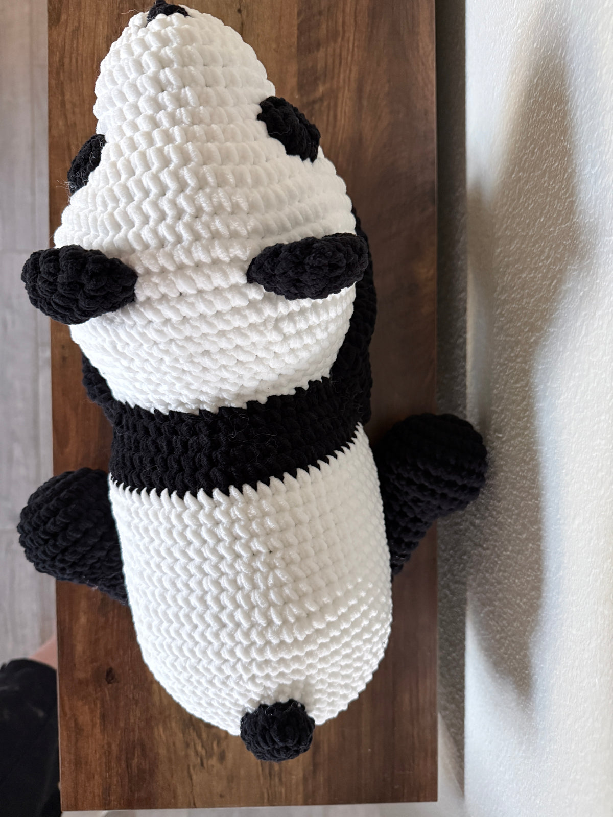 A handmade crocheted panda plush toy with a textured knit pattern, resting on a dark wooden board against a light gray wall.