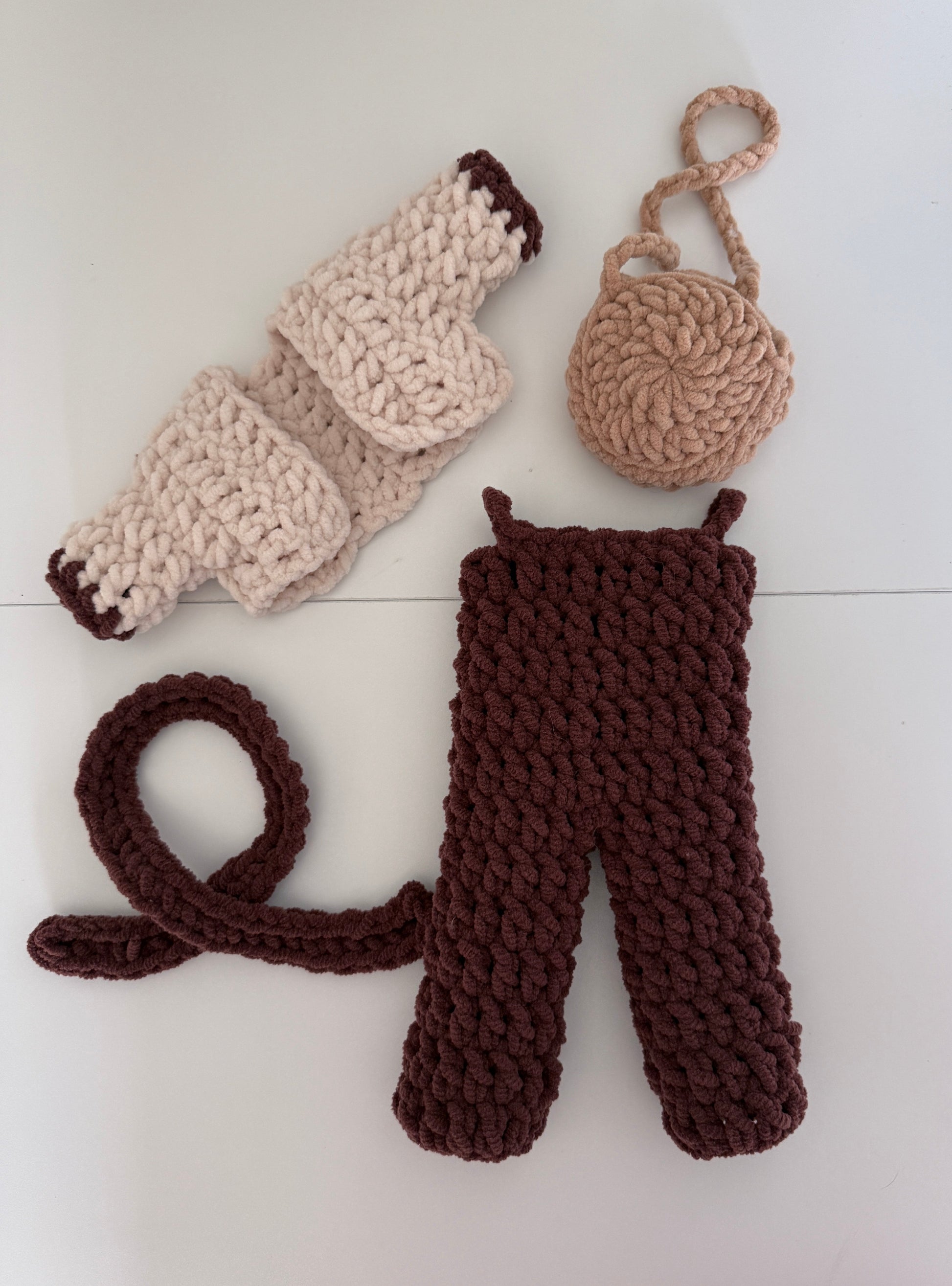 Crochet clothing items for deer including a bag, scarf, overalls, and cardigan a white surface.