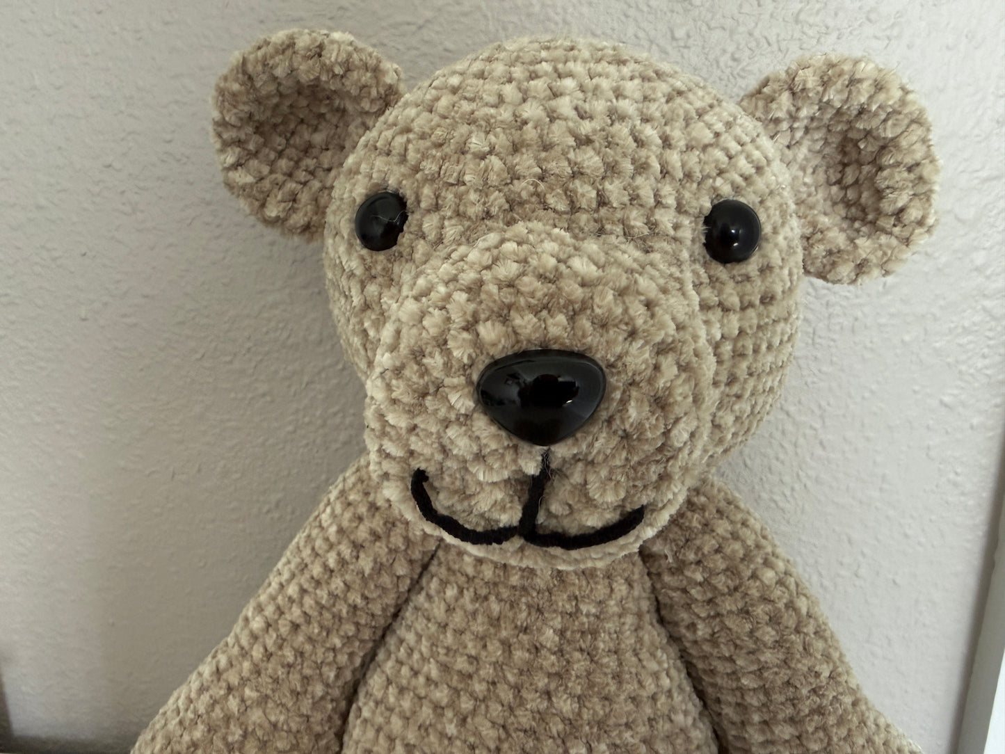 Close-up of a beige crochet teddy bear against a white wall.