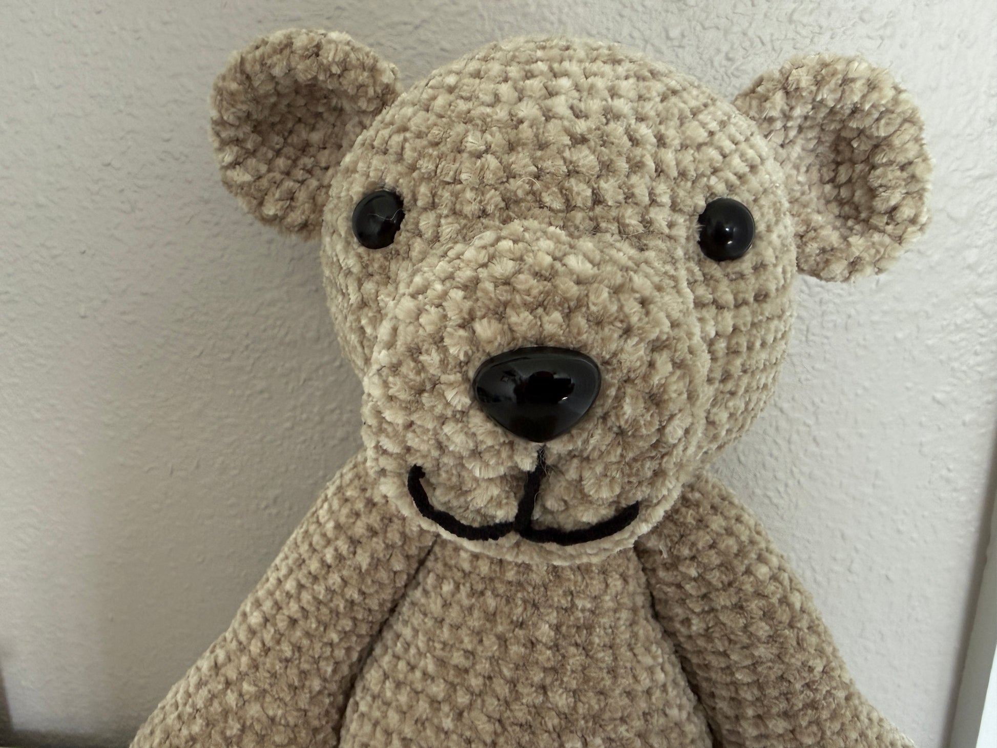 Close-up of a beige crochet teddy bear against a white wall.