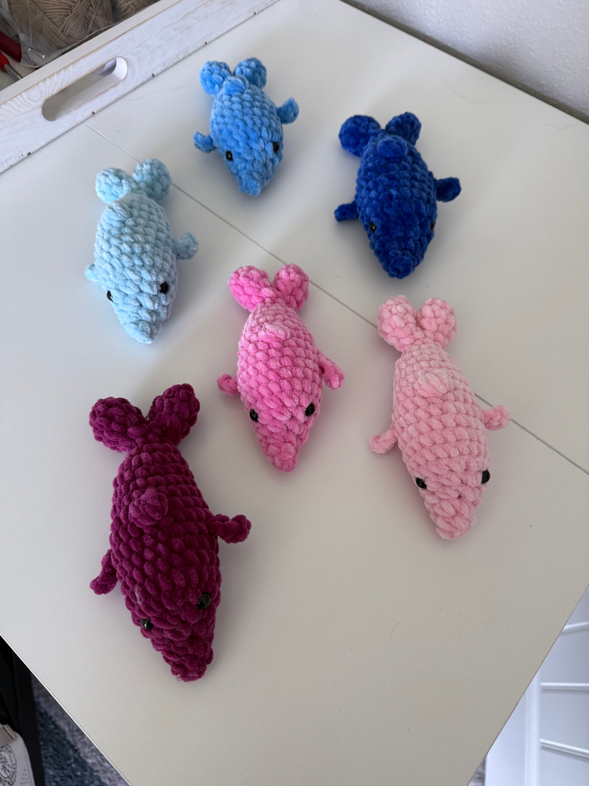 Five crocheted dolphin amigurumis in blue, pink, and purple on a white surface.